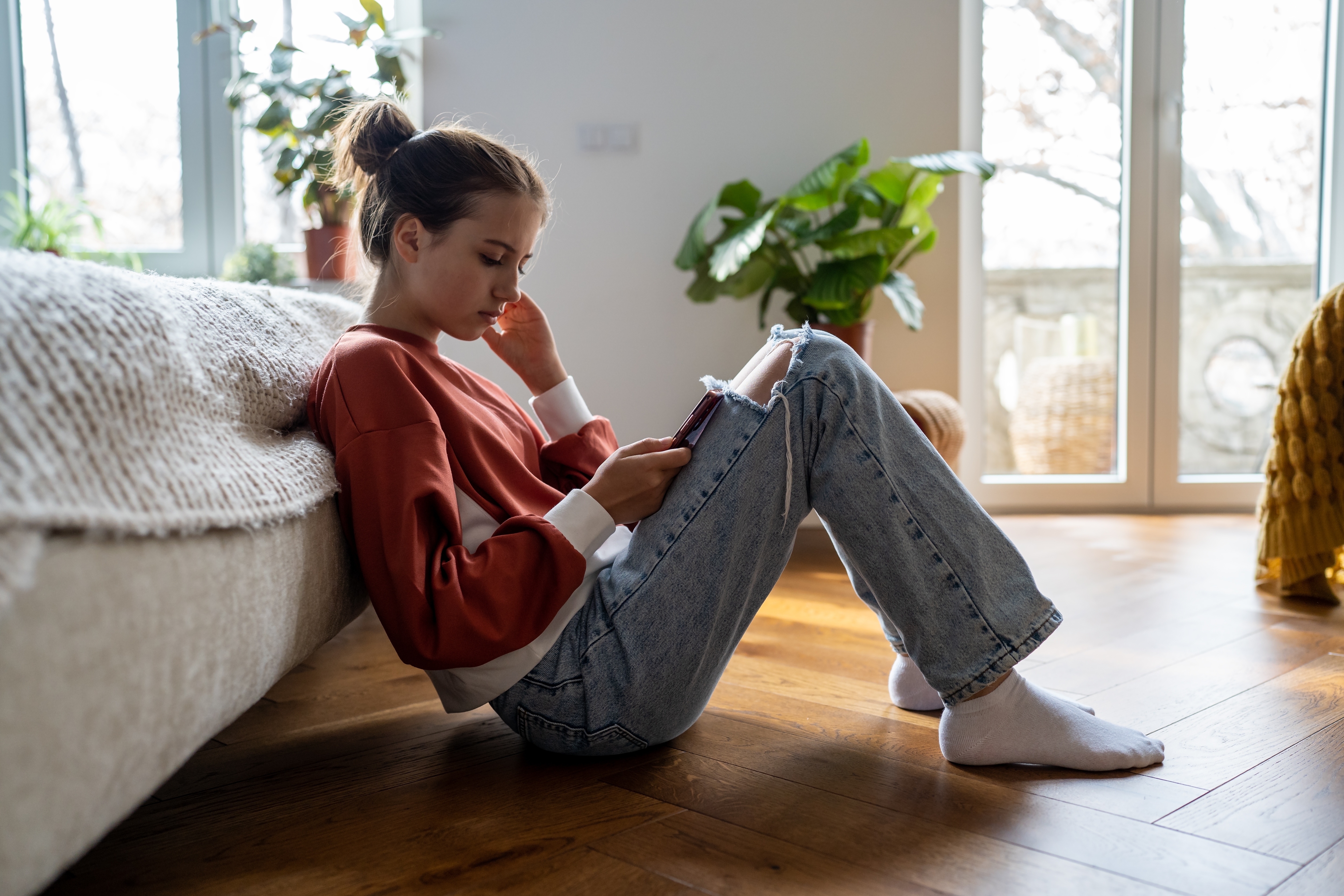 Teenage girl leaning against a couch, focused on her smartphone. She is casually dressed.